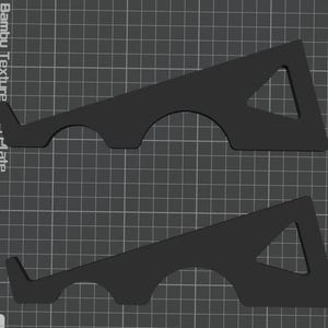 May include: Two black, geometric 3D-printed objects. Each has a triangular cutout and a curved base with a small lip. The objects are designed to hold a plate. The background is a grid.
