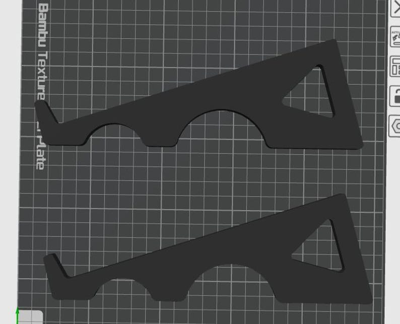 May include: Two black, geometric 3D-printed objects. Each has a triangular cutout and a curved base with a small lip. The objects are designed to hold a plate. The background is a grid.