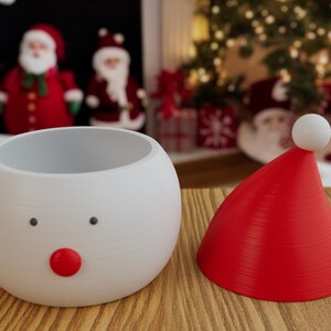 May include: A white, round, Santa-themed planter with a red nose and black eyes, next to a red Santa hat with a white pom-pom. The Christmas-themed decor is on a wooden surface, with Santa figures and a Christmas tree in the background.