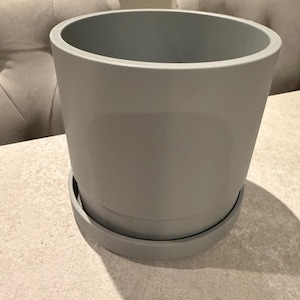 May include: A gray ceramic plant pot with a matching saucer. The pot has a cylindrical shape with a wide opening and a smooth, matte finish. The saucer is a flat, circular base that fits the pot.