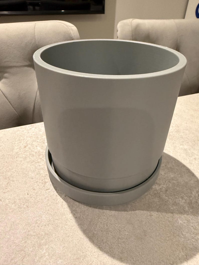 May include: A gray ceramic plant pot with a matching saucer. The pot has a cylindrical shape with a wide opening and a smooth, matte finish. The saucer is a flat, circular base that fits the pot.
