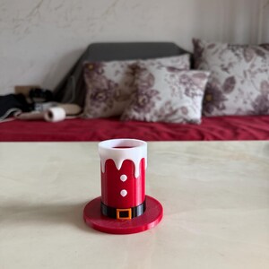 May include: A red and white Santa hat-shaped shot glass. The shot glass has a black belt with a gold buckle and two white buttons. The shot glass is sitting on a red base. The background includes a red surface and pillows.