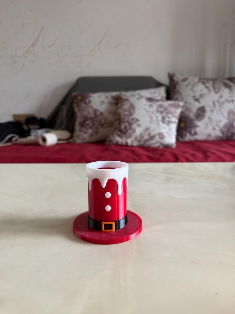 May include: A red and white Santa hat-shaped shot glass. The shot glass has a black belt with a gold buckle and two white buttons. The shot glass is sitting on a red base. The background includes a red surface and pillows.