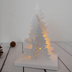 May include: A white Christmas-themed decoration featuring a cut-out design of a deer and two pine trees. The trees have star-shaped cutouts that allow light to shine through. The decoration sits on a white rectangular base.
