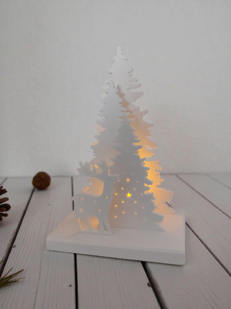 May include: A white Christmas-themed decoration featuring a cut-out design of a deer and two pine trees. The trees have star-shaped cutouts that allow light to shine through. The decoration sits on a white rectangular base.