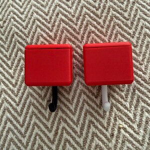 May include: Two red wall hooks with rectangular tops. One hook is black, and the other is white. The hooks are set against a textured, neutral-toned background.