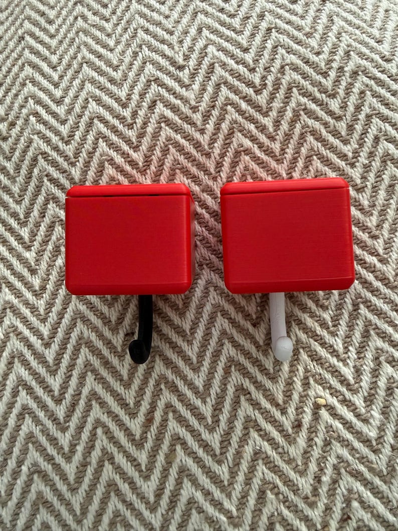 May include: Two red wall hooks with rectangular tops. One hook is black, and the other is white. The hooks are set against a textured, neutral-toned background.