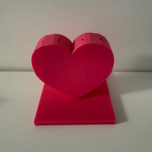 May include: A bright pink heart-shaped object with a rectangular base. The heart has a textured, ribbed design on the top portion. The object is made of a solid material and stands upright on a white surface.