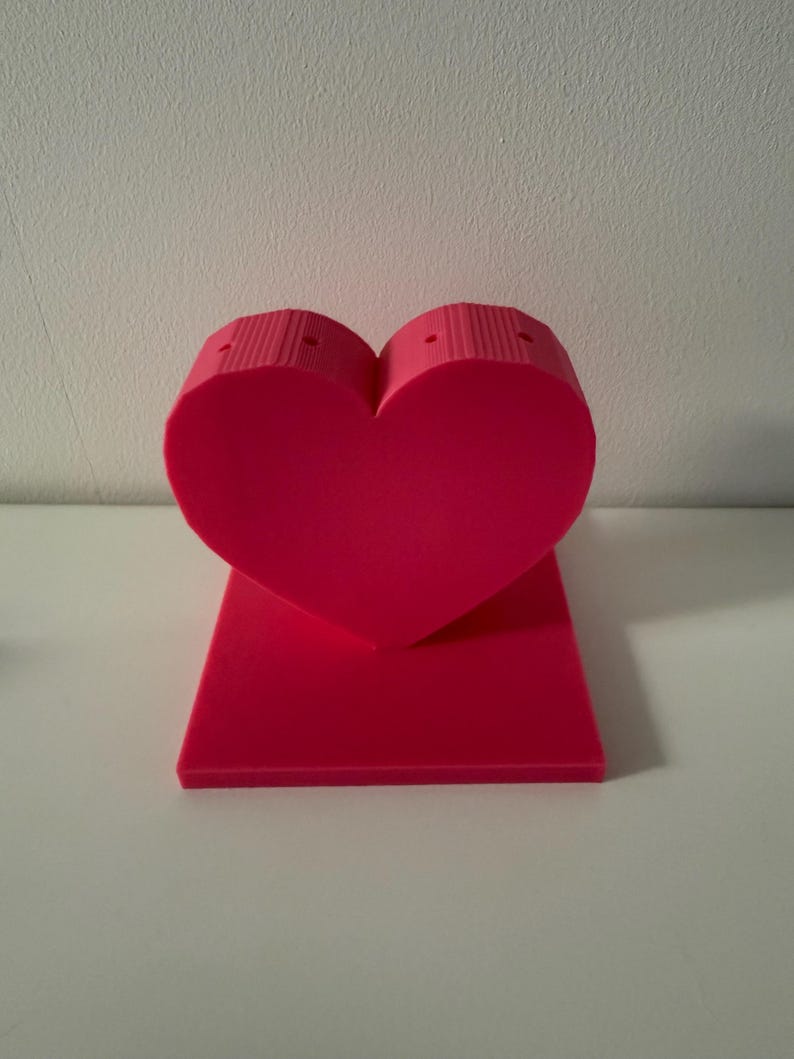 May include: A bright pink heart-shaped object with a rectangular base. The heart has a textured, ribbed design on the top portion. The object is made of a solid material and stands upright on a white surface.