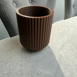 May include: A brown, cylindrical planter with vertical ribbed detailing. The top rim has a decorative, ridged edge. The planter sits on a light gray surface, with two gray upholstered chairs in the background.
