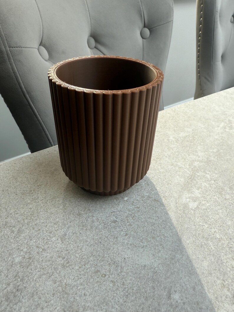 May include: A brown, cylindrical planter with vertical ribbed detailing. The top rim has a decorative, ridged edge. The planter sits on a light gray surface, with two gray upholstered chairs in the background.