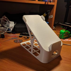 May include: A white 3D-printed phone stand with a textured surface. The stand has a rectangular base with a grid pattern and a curved top to hold a phone. The design includes a cutout for charging cables.
