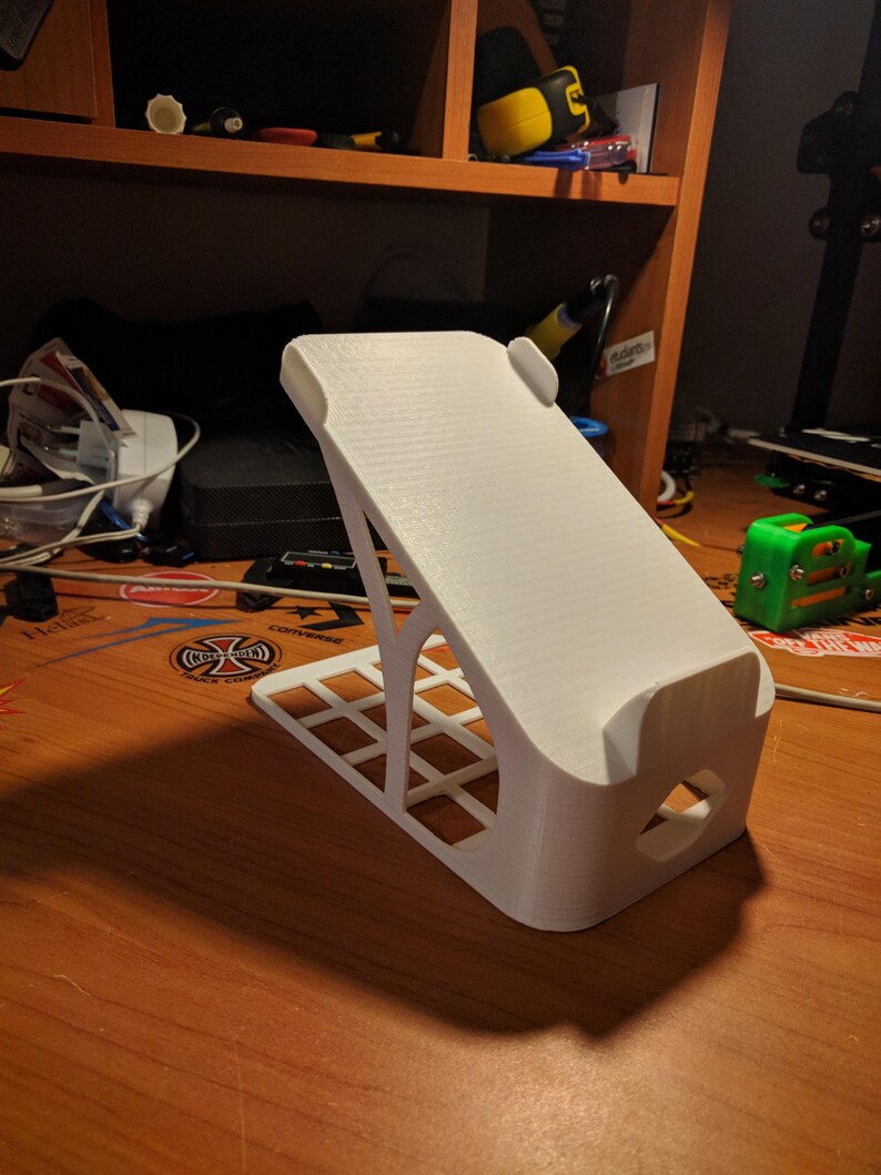 May include: A white 3D-printed phone stand with a textured surface. The stand has a rectangular base with a grid pattern and a curved top to hold a phone. The design includes a cutout for charging cables.