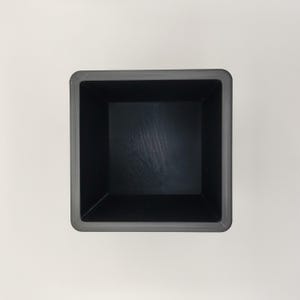 May include: A square, dark gray planter box with a textured rim and a smooth, black interior. The planter is empty and viewed from above, showcasing its simple, modern design. Ideal for small plants or succulents.