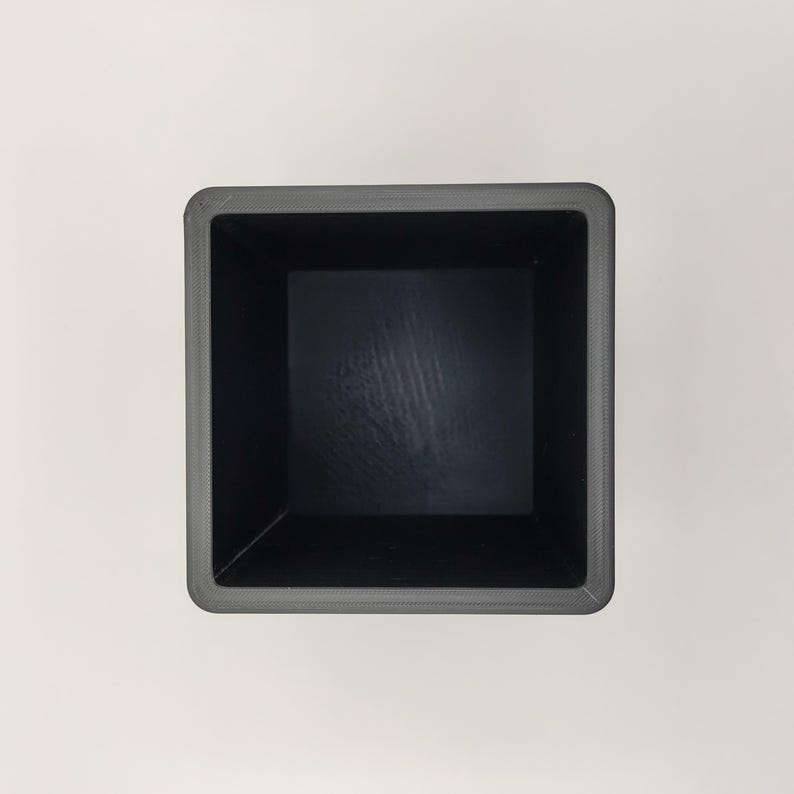 May include: A square, dark gray planter box with a textured rim and a smooth, black interior. The planter is empty and viewed from above, showcasing its simple, modern design. Ideal for small plants or succulents.