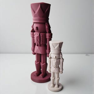 May include: Two decorative nutcrackers in a minimalist style. One is a larger, deep burgundy color, and the other is smaller and a light beige. Both have a geometric design and stand on round bases. The nutcrackers are a festive holiday decoration.