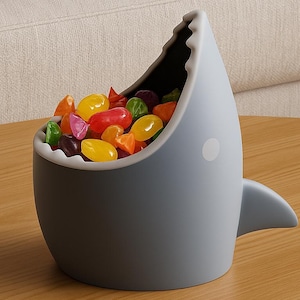 May include: A gray shark-shaped candy dish filled with colorful jelly beans. The dish has a white spot on its side and a fin. The candy dish is sitting on a wooden table.