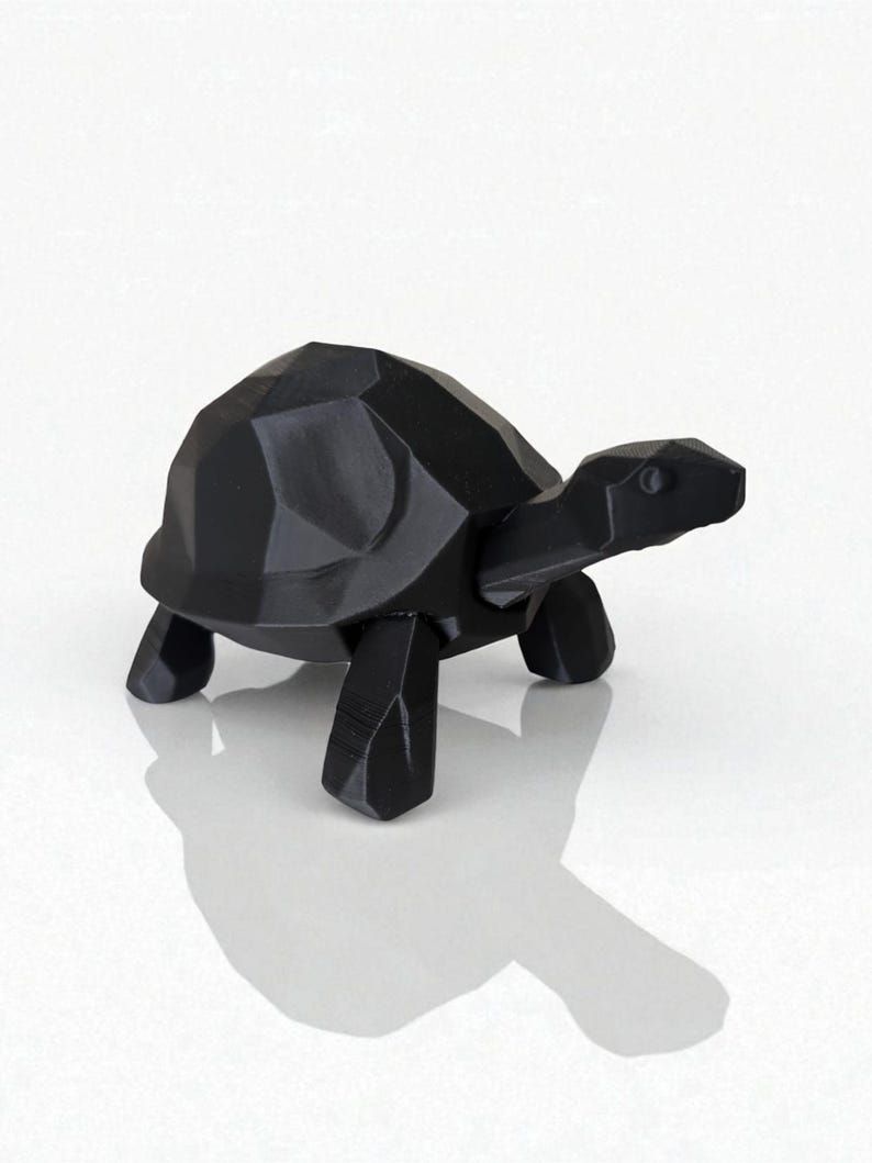 May include: A black, geometric-style turtle figurine. The sculpture has a faceted shell and a smooth, elongated head. The turtle is standing on four blocky legs. The figurine is made of a solid, dark material and casts a shadow.
