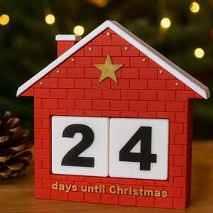 May include: A red house-shaped Christmas countdown calendar with a white roof and a gold star. The calendar displays the number 24 on white blocks, with the text "days until Christmas" below.