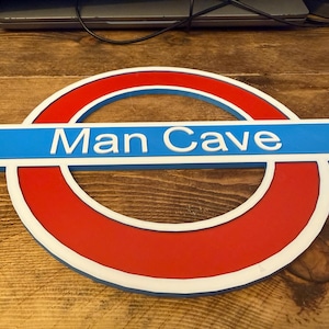 May include: A red, white, and blue sign with the text "Man Cave" in white lettering. The sign is in the shape of a circle with a horizontal rectangle across the middle. The sign is made of wood.