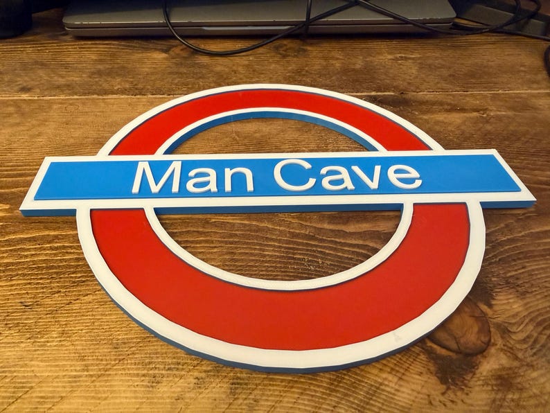 May include: A red, white, and blue sign with the text "Man Cave" in white lettering. The sign is in the shape of a circle with a horizontal rectangle across the middle. The sign is made of wood.
