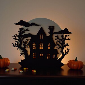 Pu&ograve; includere: Silhouette nera di una casa infestata con finestre illuminate, alberi e nuvole, ambientata contro una luna bianca. Due zucche arancioni e caramelle di mais in primo piano, creando un tema di Halloween.