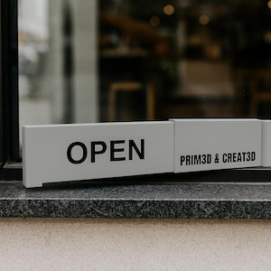 May include: A rectangular, light gray sign with the word "OPEN" in bold black letters. The sign also displays "PRIM3D & CREAT3D" in black. The sign is positioned on a dark gray stone surface, near a window.