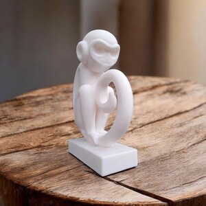Puede incluir: Una figura de mono blanco con una cola rizada, sentado sobre una base rectangular. El mono tiene una cara estilizada y est&aacute; hecho de un material liso y s&oacute;lido. La figura se exhibe sobre una superficie de madera r&uacute;stica.