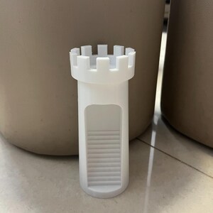 May include: A white, castle-shaped bottle stopper. The stopper has a cylindrical body with a series of steps leading to an opening. The top features a crenellated design, resembling a castle turret. The stopper is made of a solid, opaque material.