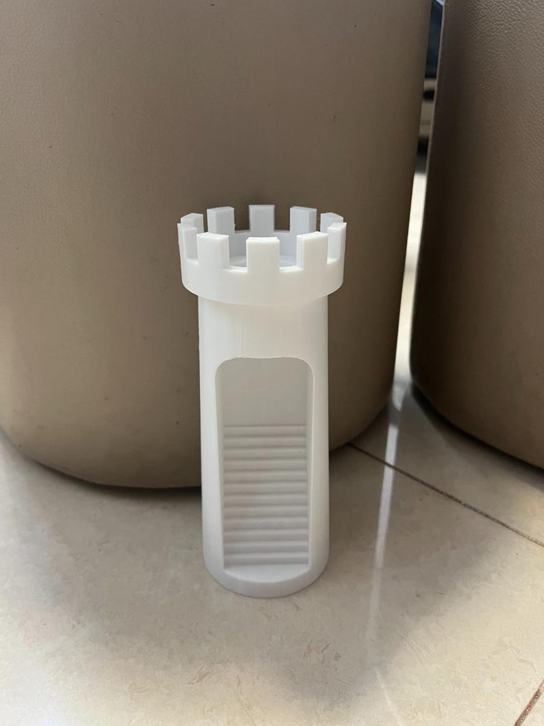 May include: A white, castle-shaped bottle stopper. The stopper has a cylindrical body with a series of steps leading to an opening. The top features a crenellated design, resembling a castle turret. The stopper is made of a solid, opaque material.
