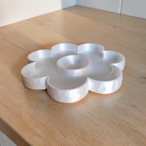 May include: A white, flower-shaped serving tray with a central dip bowl and six surrounding compartments. The tray has a pearlescent finish and sits on a wooden surface. The design is ideal for serving snacks or appetizers.