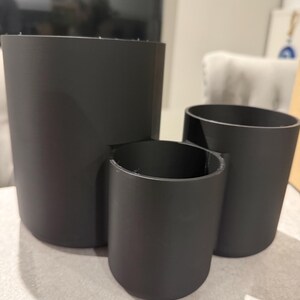 May include: Three matte black cylindrical containers of varying sizes. The largest is a tall, open cylinder. A smaller cylinder is attached to the side of the largest. The third cylinder is a smaller, standalone container.