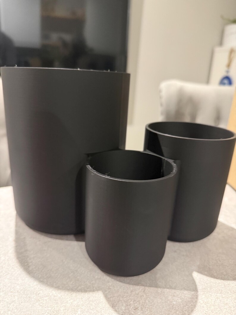 May include: Three matte black cylindrical containers of varying sizes. The largest is a tall, open cylinder. A smaller cylinder is attached to the side of the largest. The third cylinder is a smaller, standalone container.