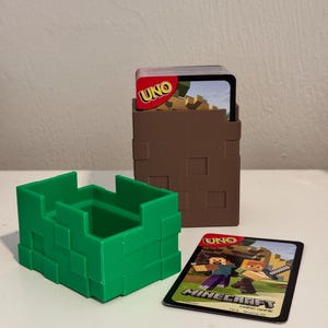 May include: A green block-shaped card holder and a brown block-shaped card holder, with a deck of UNO Minecraft cards and a single card visible. The UNO card has the Minecraft logo and characters.