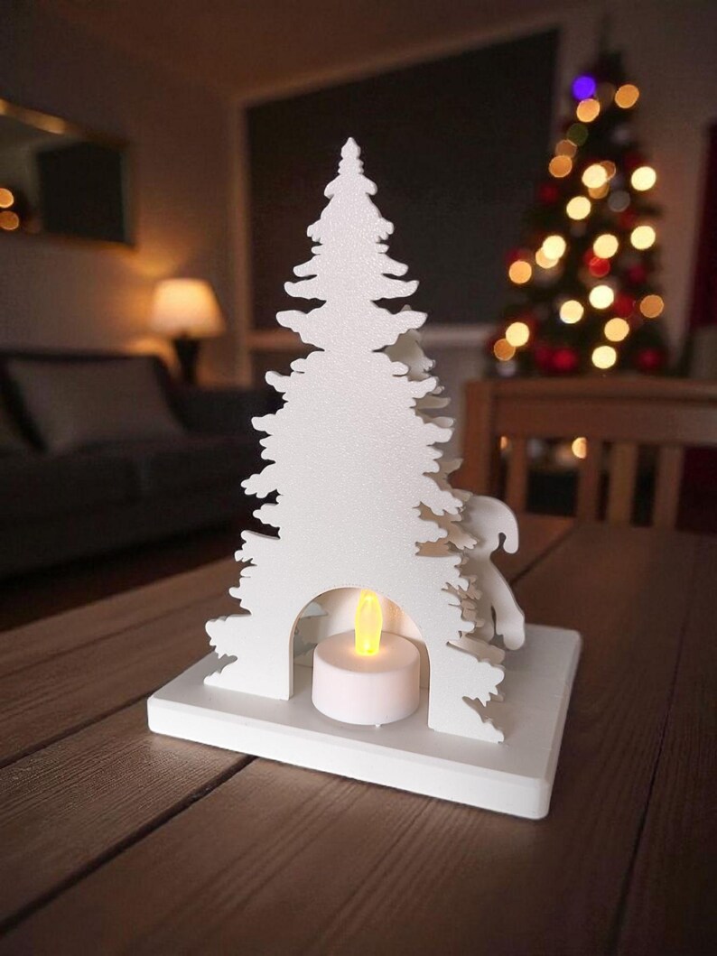 May include: A white, laser-cut Christmas tree-shaped candle holder with a small, round, battery-operated candle inside. The candle holder is on a wooden table, with a blurred Christmas tree in the background.
