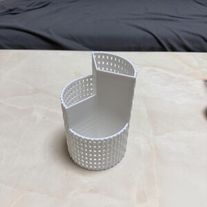 May include: A white, 3D-printed desk organizer with a unique, curved design. The organizer features a lattice-like pattern on the exterior and multiple compartments for storing pens, pencils, or other office supplies. The dimensions are approximately 4 inches wide, 4 inches long, and 4 inches tall.