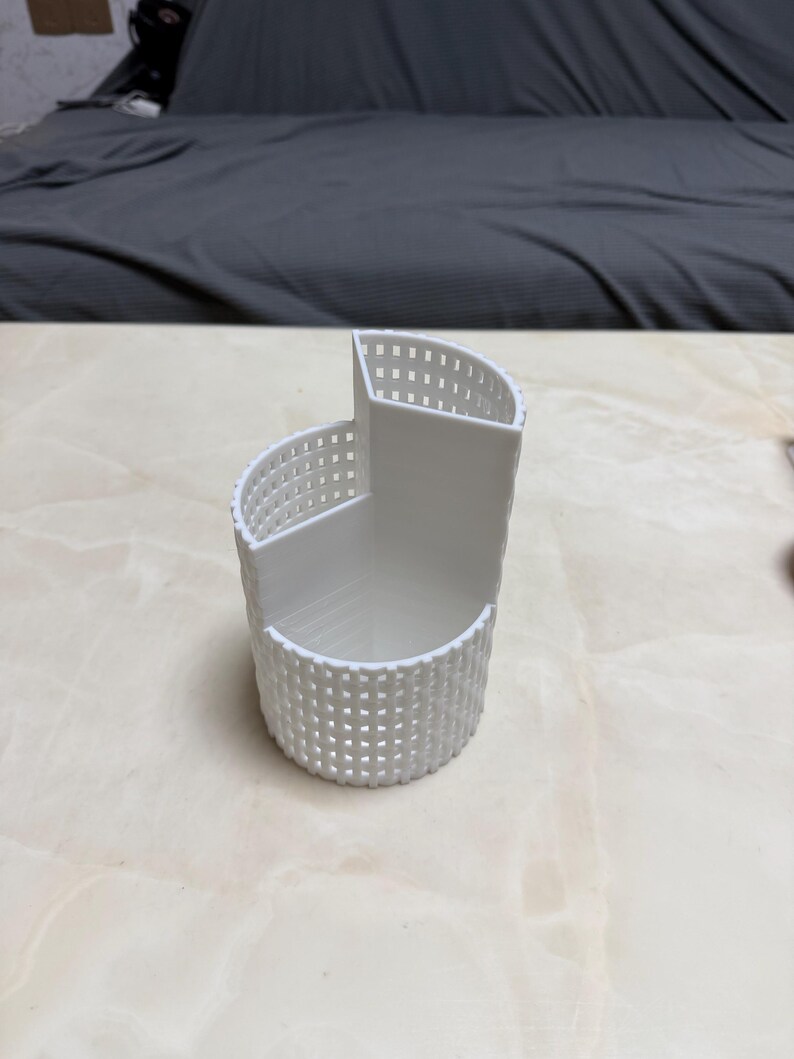May include: A white, 3D-printed desk organizer with a unique, curved design. The organizer features a lattice-like pattern on the exterior and multiple compartments for storing pens, pencils, or other office supplies. The dimensions are approximately 4 inches wide, 4 inches long, and 4 inches tall.