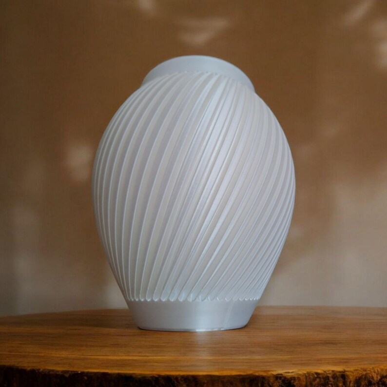 Puede incluir: Un jarr&oacute;n blanco decorativo con un dise&ntilde;o acanalado en espiral. El jarr&oacute;n tiene un cuerpo redondeado y una base ligeramente ensanchada. La superficie tiene un acabado mate. El jarr&oacute;n est&aacute; sobre una superficie de madera.