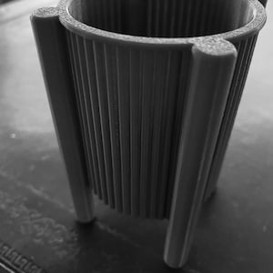 May include: A gray, 3D-printed planter with a cylindrical shape and vertical ribbed texture. The planter has three legs and a flared rim with rounded accents. The design is modern and minimalist, suitable for small plants or succulents.