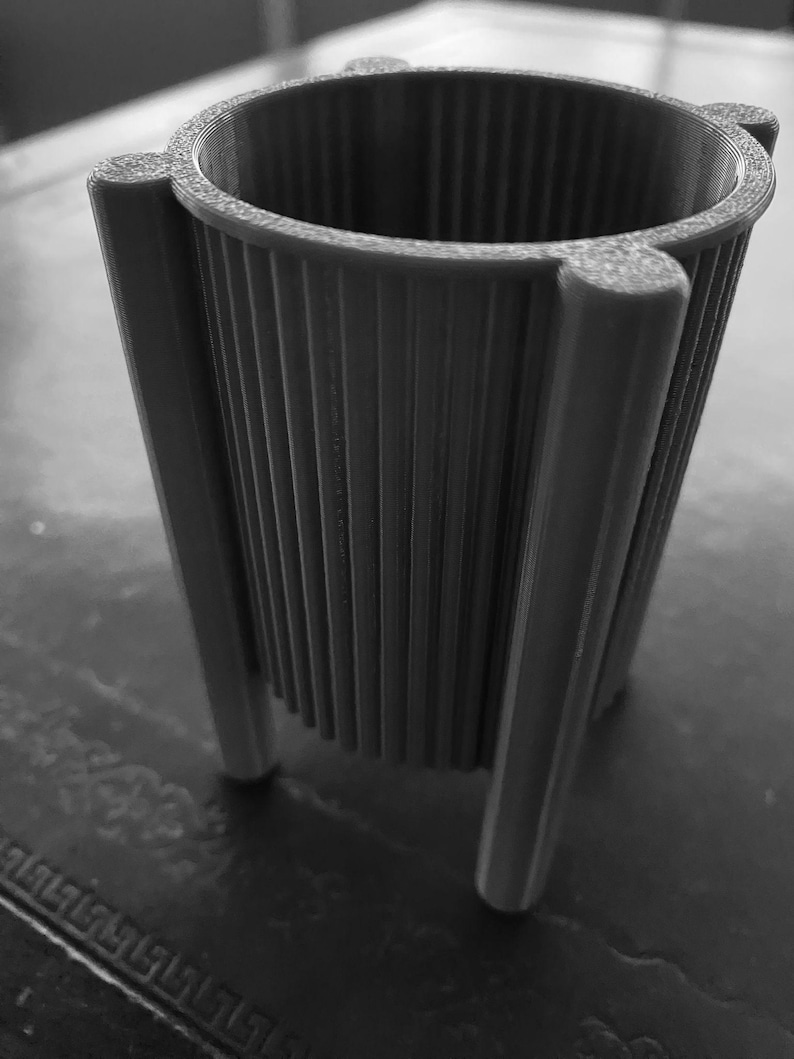 May include: A gray, 3D-printed planter with a cylindrical shape and vertical ribbed texture. The planter has three legs and a flared rim with rounded accents. The design is modern and minimalist, suitable for small plants or succulents.