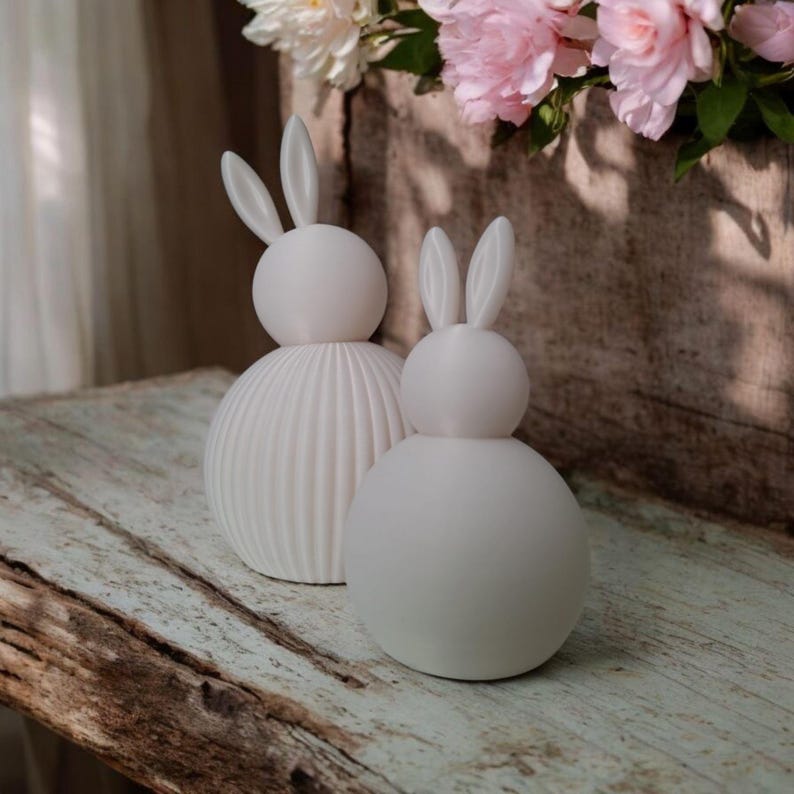 May include: Two white ceramic bunny figurines. One has a ribbed body, the other is smooth. Both have round heads and long ears. They are displayed on a distressed wooden surface, with pink and white flowers in the background.