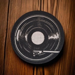 May include: A black and white vinyl record coaster with a detailed design. The coaster features concentric circles and a central white circle, with a white graphic of a record player arm. The coaster is round and sits on a wooden surface.