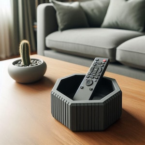 Puede incluir: Un soporte gris geom&eacute;trico para el mando a distancia con un mando dentro. El soporte est&aacute; sobre una mesa de madera, con un cactus en una maceta a la izquierda. Un sof&aacute; gris est&aacute; en el fondo.