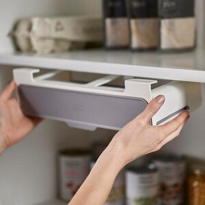 May include: A white and gray under-shelf drawer being installed in a kitchen cabinet. The drawer has a gray front and is held by a person's hands. The cabinet shelf above has a carton of eggs and spice jars.