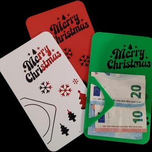 May include: Three Christmas-themed gift card holders in red, white, and green. Each card holder has the words "Merry Christmas" in a decorative font, along with snowflakes and Christmas tree designs. The green holder contains paper currency.