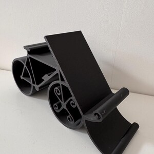 May include: Black phone stand with a bicycle design. The stand has a rectangular back and a curved base with decorative wheel-shaped elements. The stand is made of a solid black material and is designed to hold a phone or tablet.
