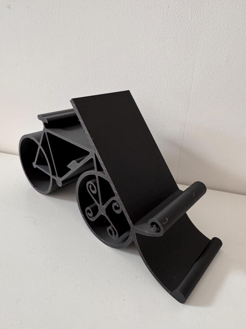 May include: Black phone stand with a bicycle design. The stand has a rectangular back and a curved base with decorative wheel-shaped elements. The stand is made of a solid black material and is designed to hold a phone or tablet.