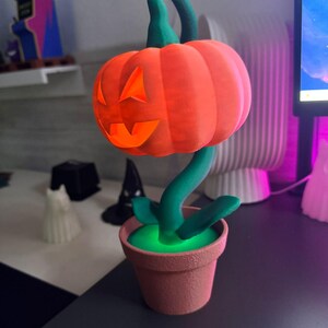 May include: A Halloween-themed decorative lamp shaped like a pumpkin with a carved face, atop a green stem and leaves, planted in a terracotta-colored pot. The pumpkin glows orange, and the pot's interior glows green.