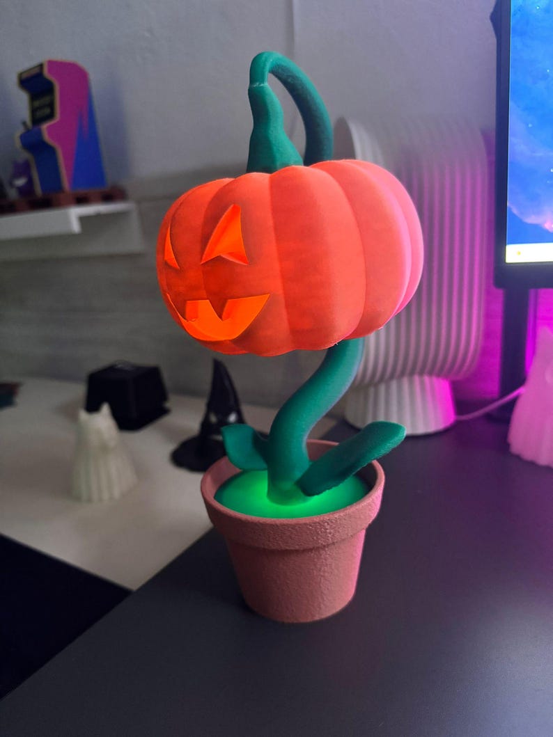 May include: A Halloween-themed decorative lamp shaped like a pumpkin with a carved face, atop a green stem and leaves, planted in a terracotta-colored pot. The pumpkin glows orange, and the pot's interior glows green.