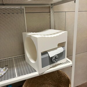 May include: A white and gray toilet paper holder with a circular opening and a slot for dispensing paper. The holder is on a white metal shelf, with a woven basket below. The design is modern and minimalist.